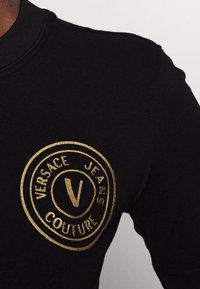 Svart bomullströja med rund guldlogotyp och texten "VERSACE JEANS COUTURE" samt en central "V"-design. Kort ärm.