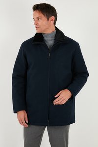 Uomo che indossa un cappotto blu navy con cerniera e collo nero, insieme a un maglione a collo alto grigio, in piedi davanti a uno sfondo bianco uniforme.