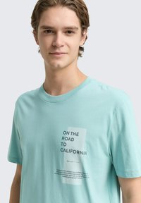 Svetlo teal bombažna majica s kratkimi rokavi, ki na sprednji strani vsebuje natisnjen tekst v moderni pisavi, obdan s kontrastnim pravokotnikom.