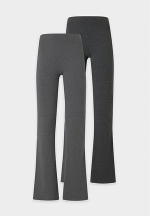 Donna con capelli intrecciati che indossa una maglietta bianca a maniche corte, pantaloni svasati grigi a vita alta e scarpe da ginnastica bianche, in piedi davanti a uno sfondo bianco semplice.