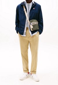 Námořnická nylonová bunda přes pruhované tričko, khaki chino kalhoty a bílé tenisky. Součástí je zelená crossbody taška s n�ápisem "TOMMY JEANS".