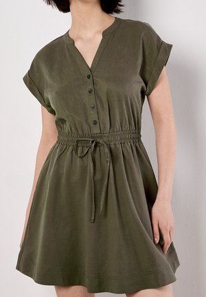 Femme portant une robe vert olive à manches courtes, avec des boutons sur le devant et une ceinture élastique nouée, devant un fond clair uni.