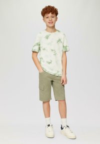 T-Shirt mit kurzen Ärmeln im Batik-Muster in Grün und Weiß, kombiniert mit olivfarbenen Cargo-Shorts. Weiße Sneakers mit marineblauen Akzenten und Crew-Socken.