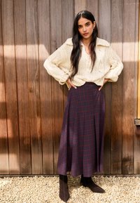 Kremasto debelo pleten pulover, temna karo midi krilo in rjavi gležnjarji. Postavljeno ob lesene obloge in prodnato tla.