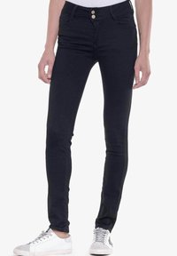 Jeans ajustados negros de mezclilla, con una cintura de altura media, dos bolsillos frontales, dos bolsillos traseros y dos botones frontales. Combinados con zapatillas blancas.