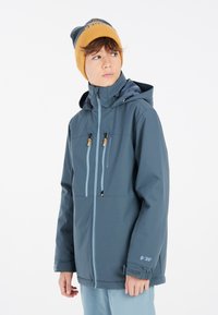 Giacca impermeabile blu con zip frontale, due tasche sul petto e cappuccio regolabile. Indossata con un berretto giallo a contrasto.