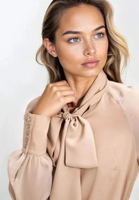Kobieta z falującymi, blond włosami, ubrana w beżową bluzkę z wiązaniem w okolicy szyi, delikatnie trzyma kokardę blisko szyi, spoglądając lekko w prawo.