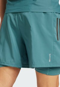 Teal sportieve shorts met een aansluitende binnenlaag, gedragen door een persoon, met in de rechteronderhoek nabij de zoom kleine tekst "CLIMACOOL".