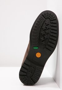 Semelle de botte en cuir marron avec une bande de roulement en caoutchouc noir robuste. Comprend des accents circulaires verts et orange ainsi qu'un logo en relief, conçu pour offrir une bonne traction.