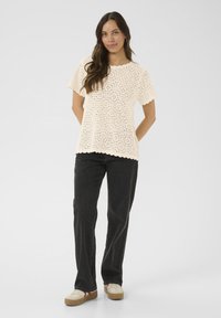 Top in crochet beige a maniche corte, con orlo a onde e motivo a rete aperta, abbinato a pantaloni neri a gamba larga e sneakers beige.