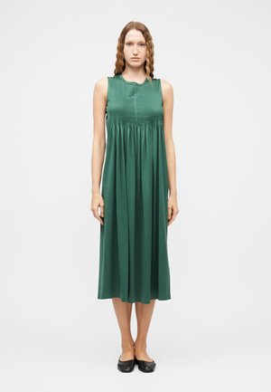 Žena s vlnitými vlasmi, oblečená v bezrukávových plisovaných tmavozelených midi šatách a čiernych balerínach, stojaca na jednoduchom svetlom pozadí.