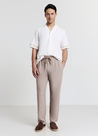 Weißes Kurzarmhemd mit strukturierten Manschetten, kombiniert mit hellbraunen, locker sitzenden Hosen mit Taschen und einem Taillenzug. Braune Espadrilles.