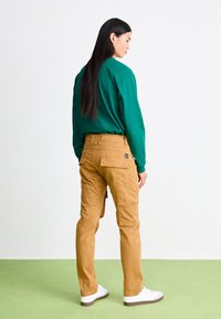 Cargo-Hosen in Beige mit Seitentaschen, kombiniert mit einem grünen Langarmshirt. Das Modell trägt weiße Sneakers mit braunen Sohlen.