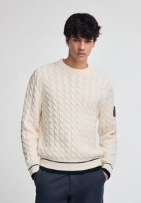 Kabelstrick, Cremefarbener Pullover mit Rundhals, gerippten Bündchen und Saum. Verfügt über einen marineblauen Streifen am Saum. Beinhaltet ein Patch am Ärmel.