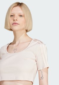 Světle růžový zkrácený top s žebrovanou texturou, kulatým výstřihem a krátkými rukávy, které mají jemné vodorovné pruhy a malý detail loga.