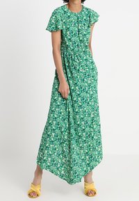 Grön blommig maxiklänning med korta volangärmar, rynkad midja och flödande nederdel. Styld med gula sandaler.
