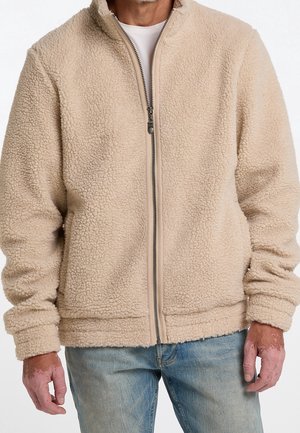 Man wearing beige fleece zip-up jacket over white shirt and light blue jeans, standing with arms relaxed by sides.