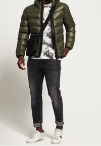 Chaqueta acolchada verde oliva con capucha, camiseta blanca con gráfico, vaqueros oscuros y zapatillas blancas. Lleva un bolso negro de bandolera.