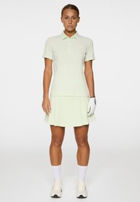 Helles grünes Golf-Outfit mit einem kurzärmligen Polo und einem plissierten Rock. Das Material wirkt glatt mit subtiler Textur. Weiße Handschuhe und Schuhe.