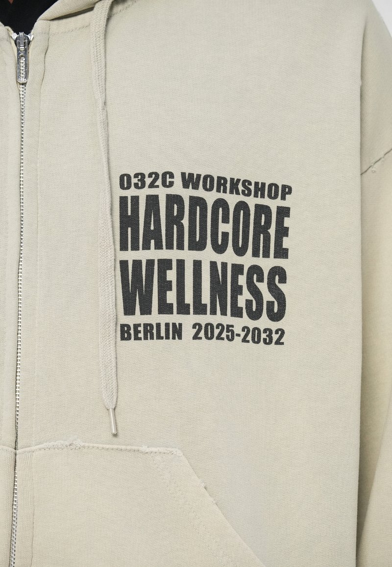 Lichtbeige rits hoodie met zwarte tekst "032C WORKSHOP HARDCORE WELLNESS BERLIN 2025-2032" op de borst boven de zak.