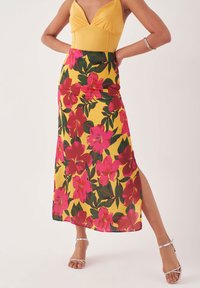 Florale maxi rok in geel met grote roze bloemen en groene bladeren, voorzien van een zijopening en soepel, lichtgewicht materiaal; gecombineerd met een gele top.