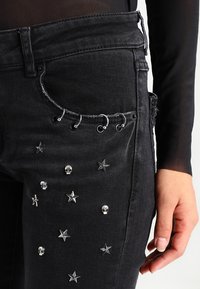 Černé džíny z denimu se stříbrnými hvězdami a lebkami s cvočky, malé stříbrné kroužky na okraji přední kapsy, nošeny s průhledným černým topem s dlouhým rukávem.