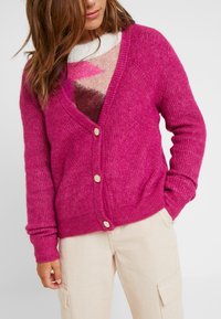 Femme portant un cardigan tricoté rose vif avec des boutons dorés par-dessus un pull multicolore et un pantalon cargo beige, main dans la poche.