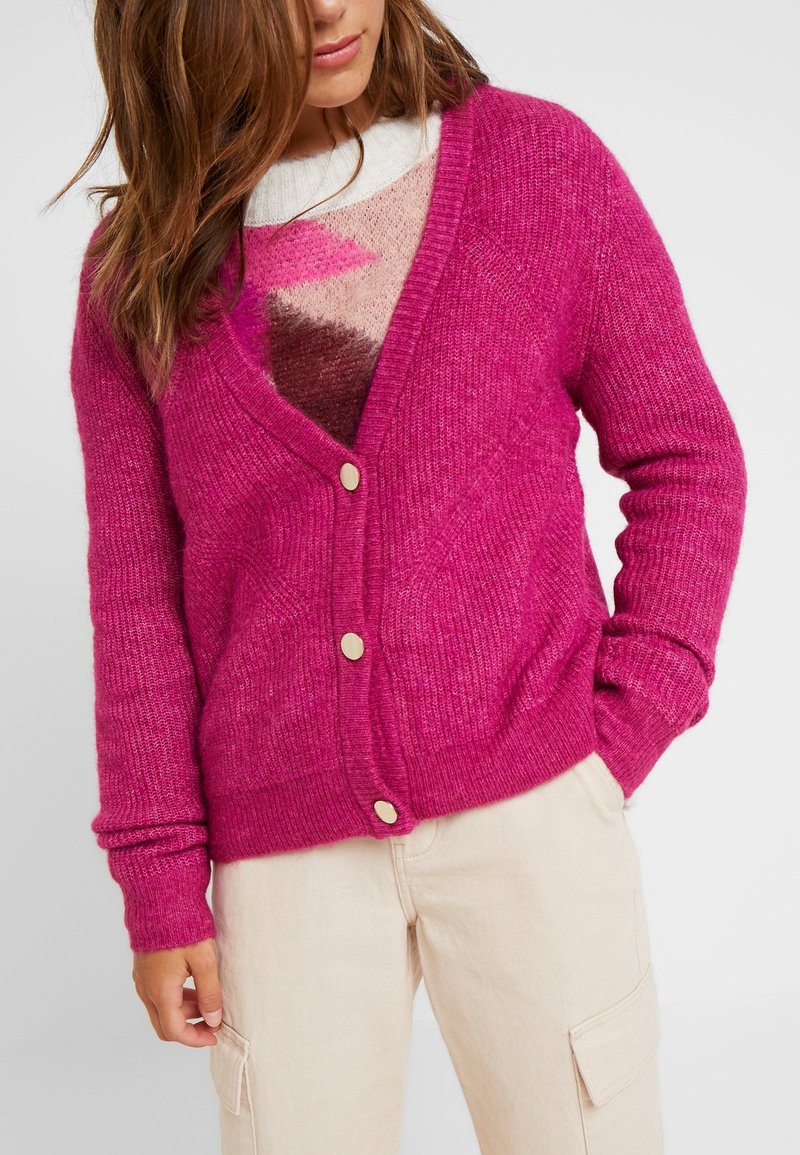 Femme portant un cardigan tricoté rose vif avec des boutons dorés par-dessus un pull multicolore et un pantalon cargo beige, main dans la poche.