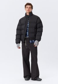 Ung mand iført en sort puffjakke, blåt ternet skjorte, løse sorte bukser, grå fingerløse handsker og sorte sneakers, stående foran en hvid baggrund.