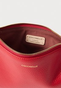 Piros bőr tasak apró szemcsés textúrával, arany cipzárral és belső szövetbéléssel. A címkén az szerepel: "Coccinelle Eredeti Olasz Bőr."