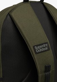 Correa de mochila acolchada de color verde oliva con respaldo de malla negra y parche con el logo "Superdry Outdoor".
