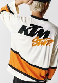 Bijela košulja dugih rukava s crnim "KTM" logotipom i narančastim mrežnim panelom s crnim prugama. Glatka tekstura, sportski dizajn.