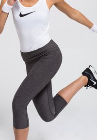 Haut blanc sans manches avec un logo Nike noir, associé à des leggings capri gris foncé. Chaussures de sport noires avec des accents blancs.