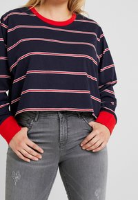 Granatowa bluzka w paski z długim rękawem, z czerwonymi mankietami i dekoltem, połączona z szarymi dżinsami z delikatnymi zdobieniami. Casualowy, wygodny krój.