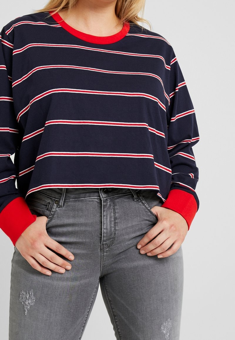 Granatowa bluzka w paski z długim rękawem, z czerwonymi mankietami i dekoltem, połączona z szarymi dżinsami z delikatnymi zdobieniami. Casualowy, wygodny krój.