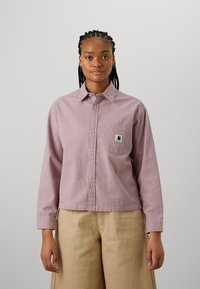 Långärmad, rosa texturerad skjorta med knäppning fram, som har en bröstficka och logopatch. Bärs tillsammans med ljusbeige byxor.