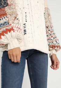 Silný pletený svetr v krémové barvě s barevnými rukávy s motivem v červených, modrých a béžových tónech; žebrované manžety a spodní lem.