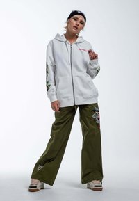 Sudadera blanca con cremallera y diseño bordado en verde y rojo, combinada con pantalones cargo holgados en verde oliva con parches multicromáticos.