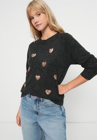 Ciemnoszary sweter dzianinowy z metalicznymi różowymi złotymi wzorami w kształcie serca. Długie rękawy i okrągły dekolt, w połączeniu z jasnoniebieskimi dżinsami.