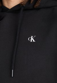 Sudadera negra de tela suave con cordón. Presenta un pequeño logo blanco "ck" en el pecho. Textura lisa y diseño ajustado.