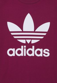 Bordeaux t-shirt med en stor vit Adidas trefoil-logotyp och varumärkesnamnet "adidas" visat nedan i fet, blockbokstäver. Slät textur.