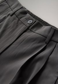 Grijze plissébroek met een gladde stoffen textuur. Voorzien van een zwarte knoop en een elastische tailleband voor een verstelbare pasvorm.