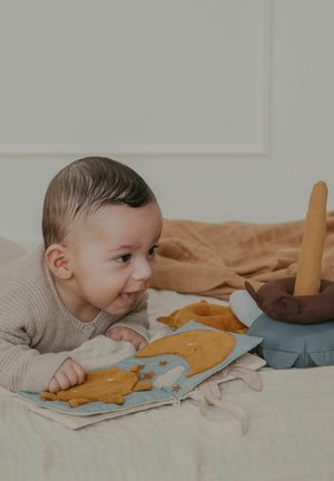 Baby ligger på maven og leger med en blød stofbog og et blødt stabledyr på en beige sengetæppe.