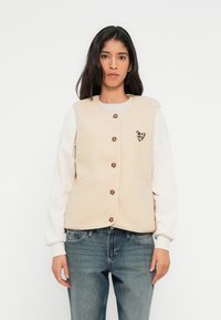 Μπεζ φλις γιλέκο με κουμπιά μπροστά, με μικρή καρδιά σε λεοπάρ μοτίβο, φορεμένο πάνω από ένα ανοιχτόχρωμο μακρυμάνικο πουκάμισο και μπλε τζιν.