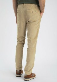 Homme portant un pantalon en velours côtelé beige et des baskets marron, se tenant les mains dans les poches, vu de dos sur un fond uni.