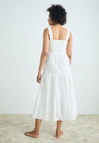 Balta medvilninė suknelė su surištu liemeniu, platesnėmis petnešėlėmis, sluoksniuota sijonu ir iki vidurio blauzdos ilgio. Lygus audinys be raštų ar akcentų.