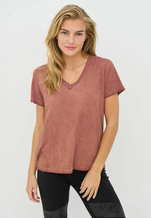 Lyshåret kvinde iført en falmet rosenfarvet V-hals kortærmet skjorte og sorte bukser, stående mod en ensfarvet hvid baggrund.