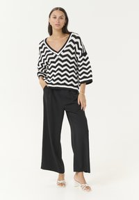 Pull en tricot à motif chevrons noir et blanc avec un décolleté en V profond, associé à un pantalon noir ample et des sandales à talons blanches.