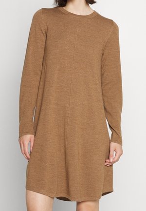 Kvinde iført en langærmet, knælang, kamelbrun strikket kjole med rund hals mod en ensfarvet lys baggrund.
