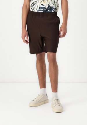 Braune gerippte Shorts mit einem elastischen Bund, seitlichen Taschen, getragen mit einem gemusterten Hemd und cremefarbenen Sneakers vor einem weißen Hintergrund.